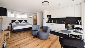 A modern bedroom with a bed, bedside tables, a desk, and a chair. The room has wooden flooring and a large window.