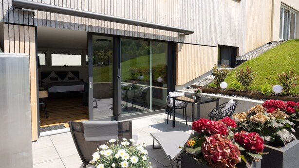 A modern house with a glass door entrance, a patio with a table and chairs, and a garden with flowers.