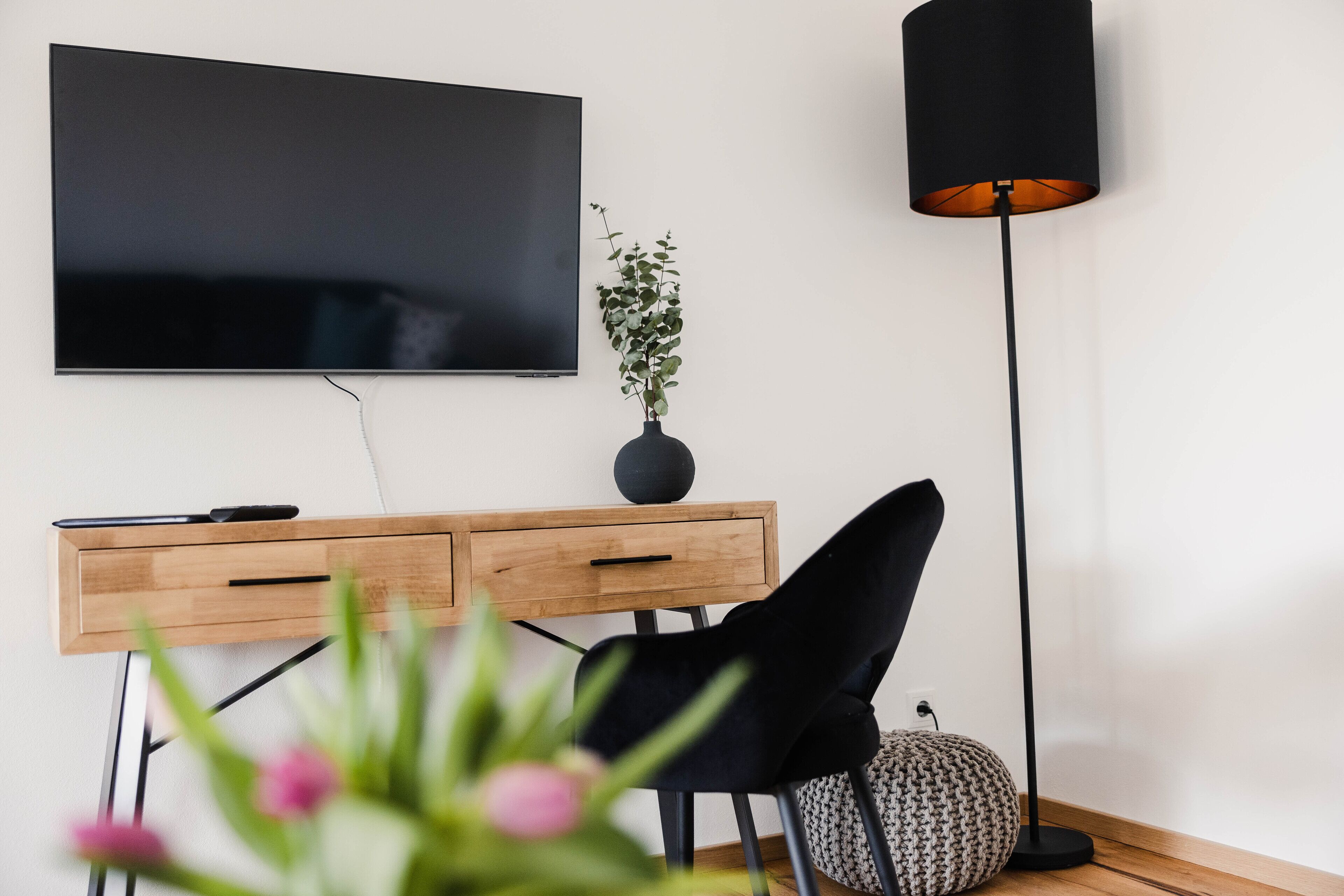 A modern living room with a flat-screen TV mounted on the wall, a wooden side table with a vase of green plants, a black chair, and a tall floor lamp.