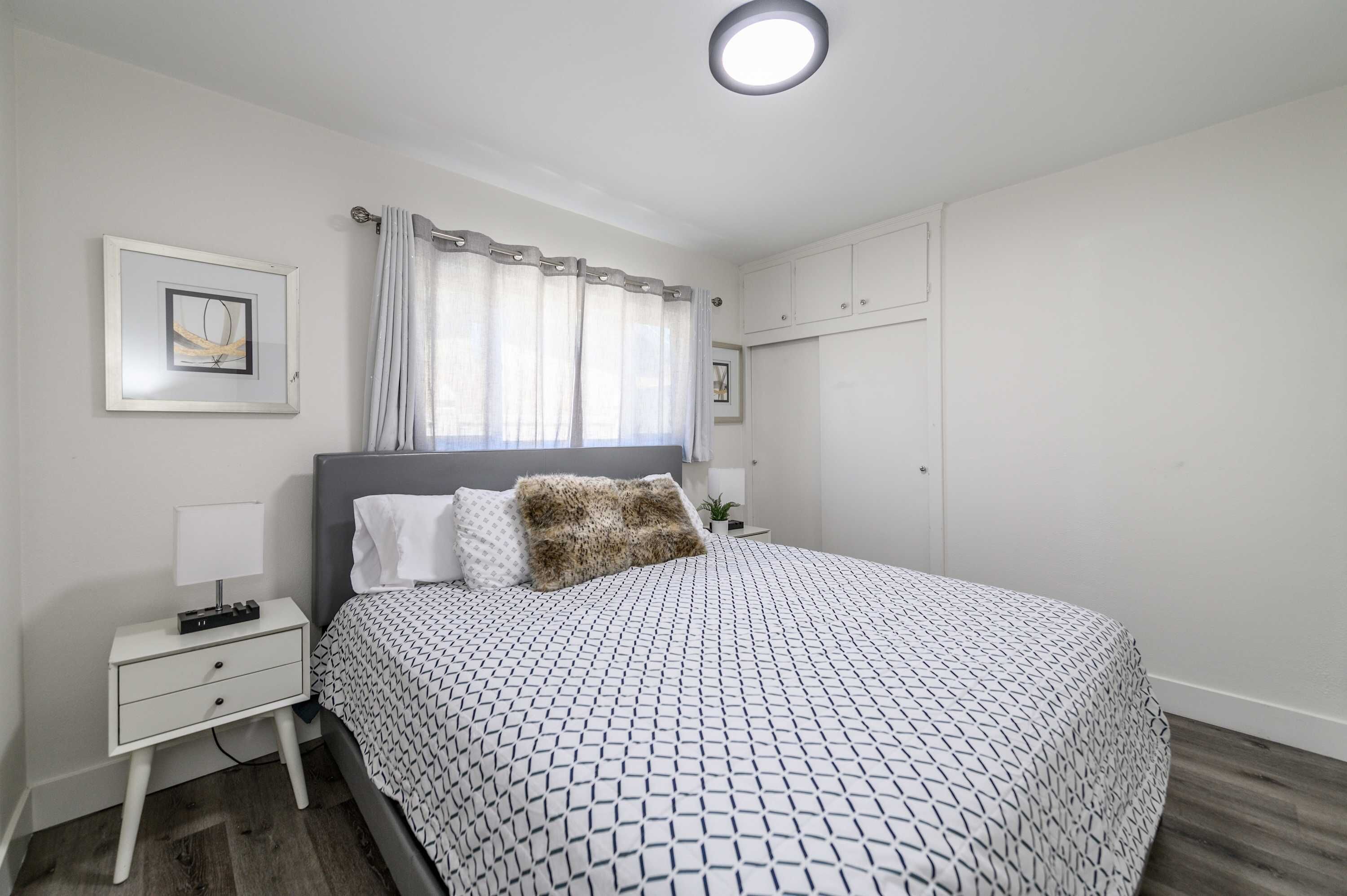 A bedroom with a bed, a nightstand, a bedside table, a window with curtains, and a ceiling light.