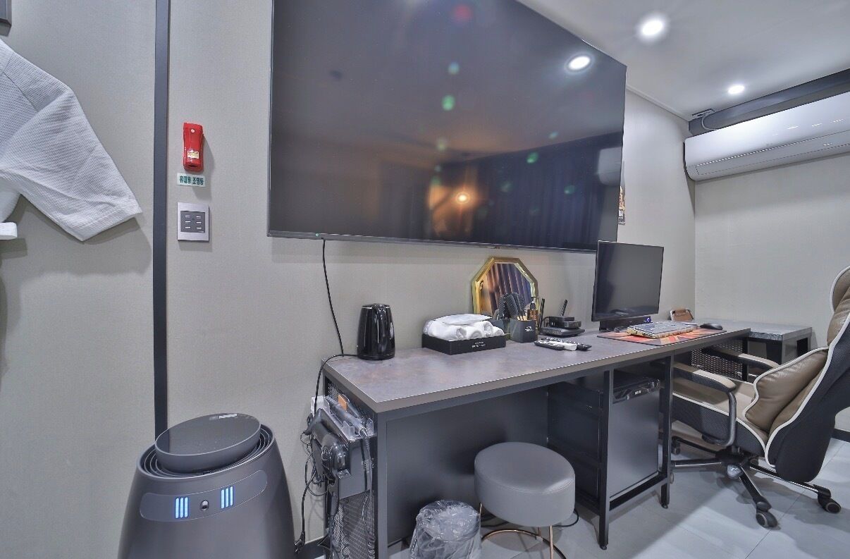 A modern office with a large flat-screen TV, a desk with a computer, a chair, and a trash bin.