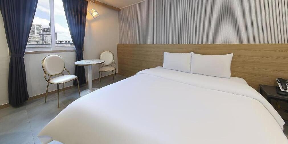 A hotel room with a large bed, a small round table, two chairs, a nightstand, and a view of the city through the window.