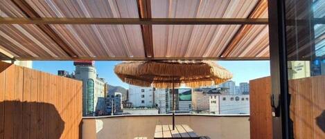 Una terraza en la azotea con una mesa y bancos de madera, un toldo de paja y vistas a la ciudad.