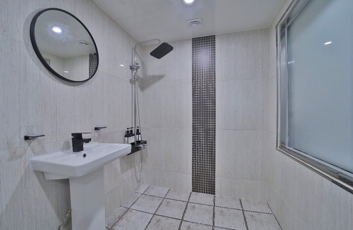 Een badkamer met een witte wastafel, een ronde spiegel, een douche met een zwarte douchetuit en betegelde wanden.