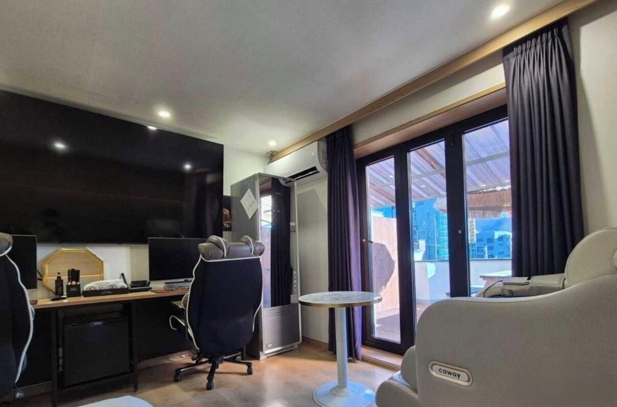 Quarto de hotel com uma mesa, duas cadeiras, televisão, uma mesinha e uma janela grande com cortinas.