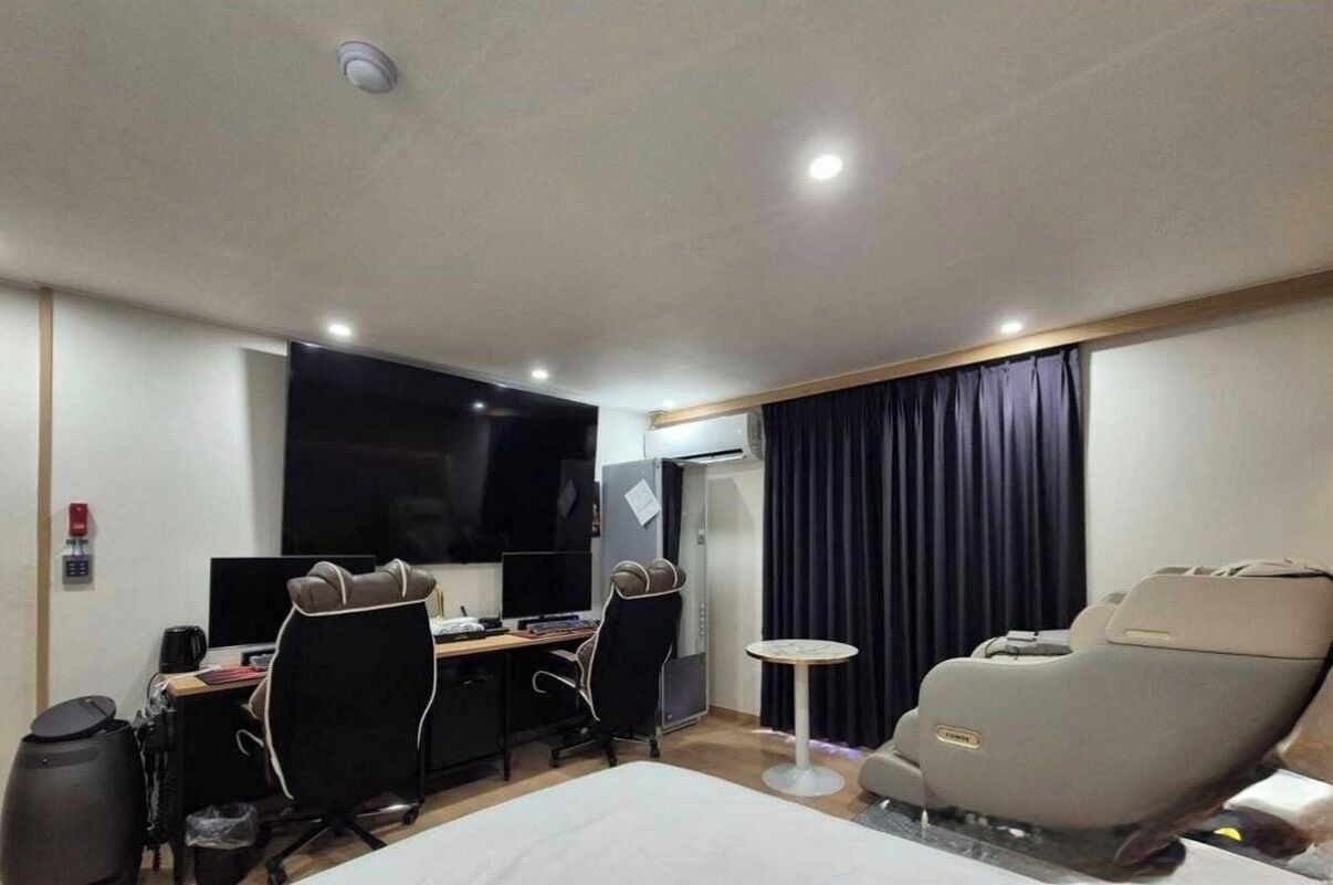 A hotel room with a bed, two chairs, a desk with a computer, a TV, a massage chair, and a small table.