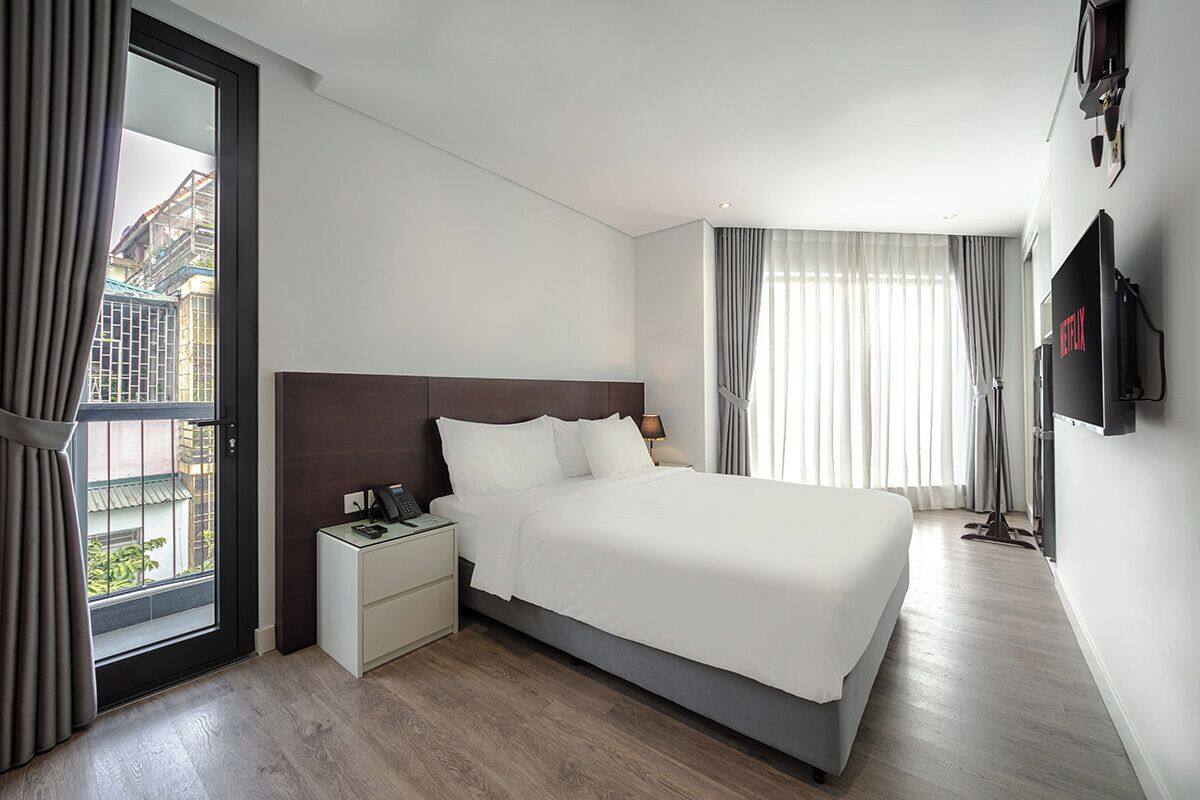 A hotel room with a bed, bedside table, lamp, TV, and a view through the window.