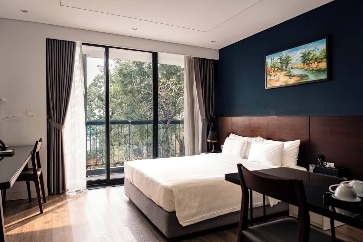 A hotel room with a bed, a desk, a chair, a nightstand, and a painting on the wall.