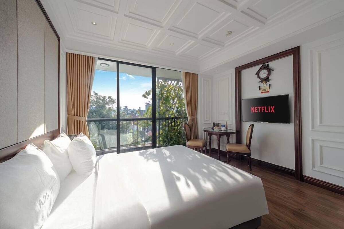 A hotel room with a large bed, a TV mounted on the wall, a balcony with curtains, and a clock above the TV.