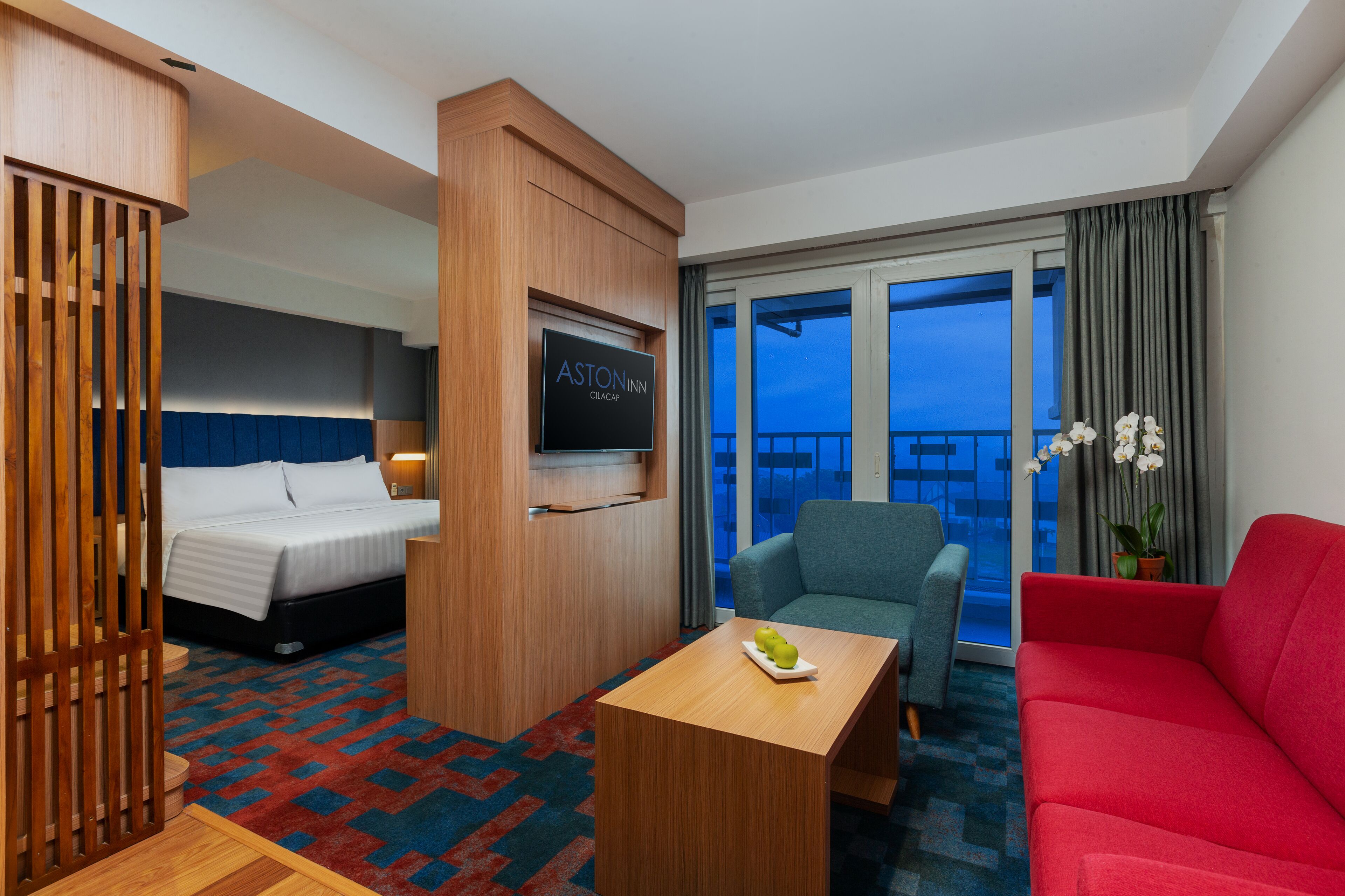 Ein Hotelzimmer mit Bett, Schreibtisch, einem roten Sofa, einem blauen Sessel und Blick auf die Stadt.