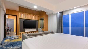 Habitación de hotel con una cama grande, televisión, zona de estar con sofá y sillones, y balcón con vistas.