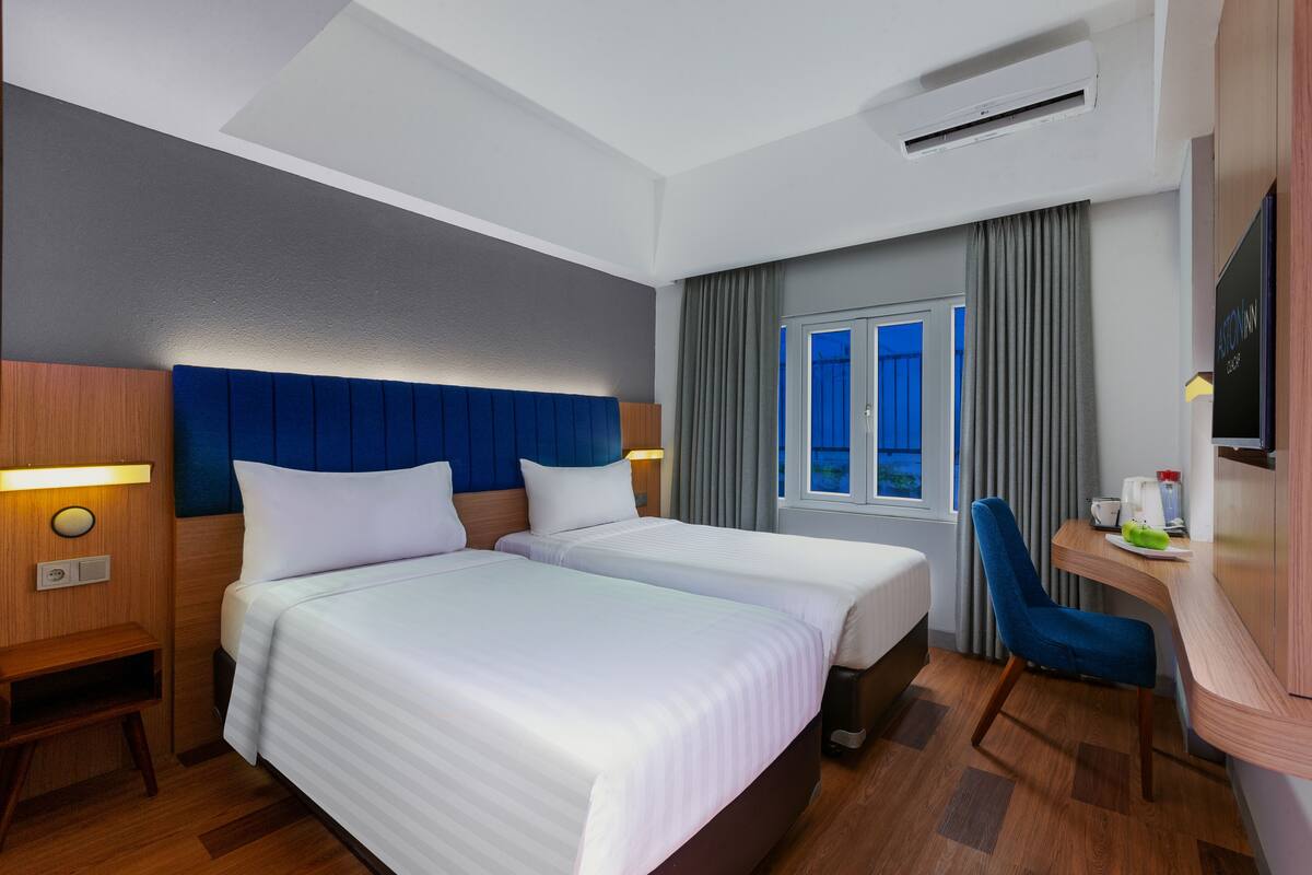 A hotel room with two beds, a desk, a chair, a TV, and a window with curtains.