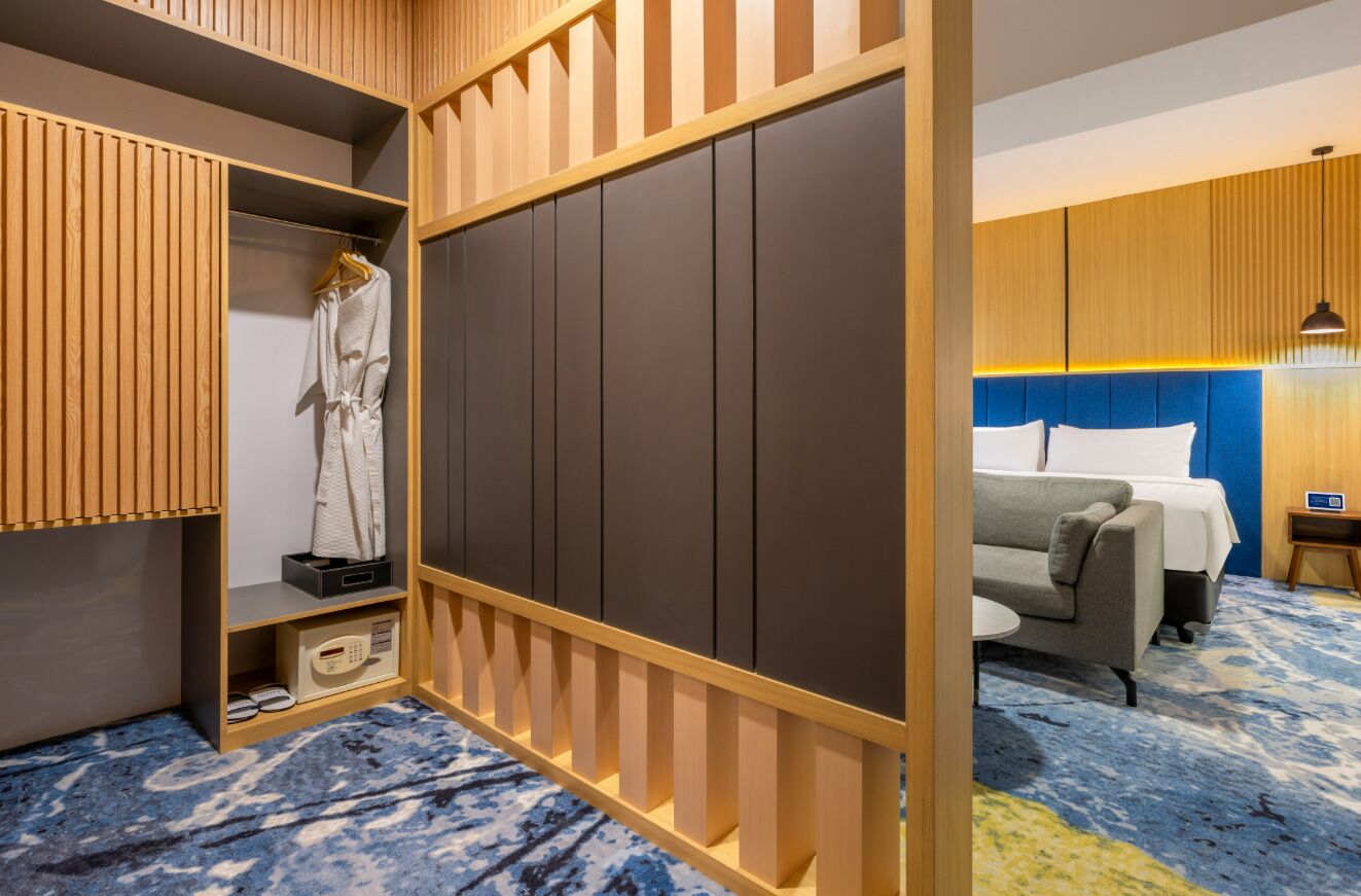 A hotel room with a wooden wardrobe, a bed with a blue headboard, a grey sofa, and a blue patterned carpet.