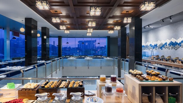 Un restaurante moderno con vistas a la ciudad, que ofrece un bufé variado y un área de estar con mesas y sillas.