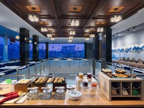 A modern restaurant with a city view, featuring a buffet with various food items and a seating area with tables and chairs.