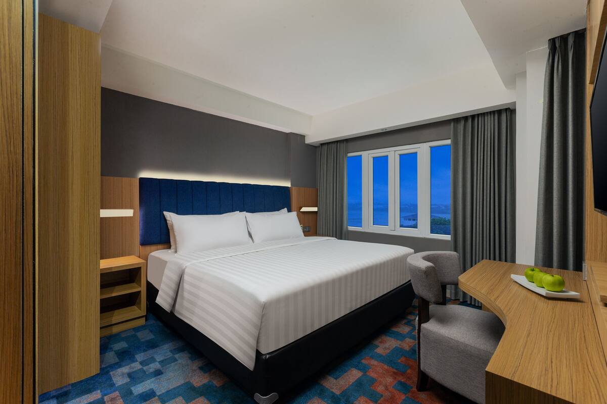 A hotel room with a bed, a desk, a chair, a window with curtains, and a view of the outside.