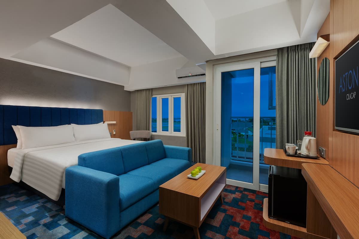 A hotel room with a bed, a blue sofa, a small table with a plate of apples, a TV, and a balcony with curtains.