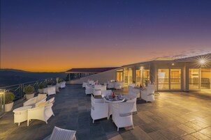 A rooftop terrace with white wicker furniture and potted plants, overlooking a cityscape at dusk.