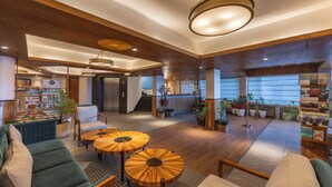 Un vestíbulo amplio con piso de madera, una recepción y una escalera que conduce a un piso superior. Hay varias zonas de estar con mesas redondas y una variedad de plantas.