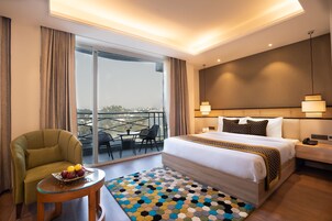 A hotel room with a large bed, a small table with fruit, a view of the outdoors through a sliding door, and a balcony with a table and chairs.