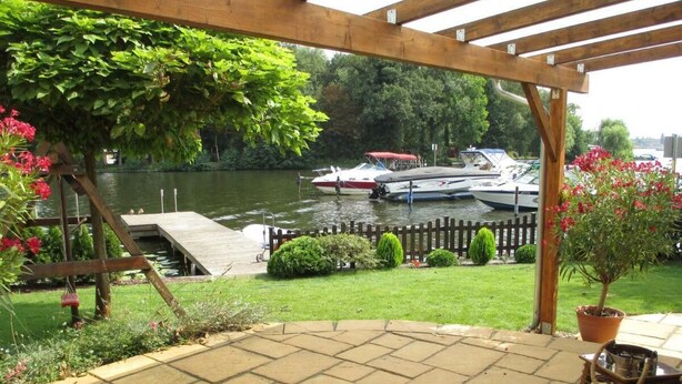 A covered patio with a view of boats on a lake, a wooden deck, and potted plants. - Directly on the water with its own jetty in Berlin - Köpenick (Berlin)