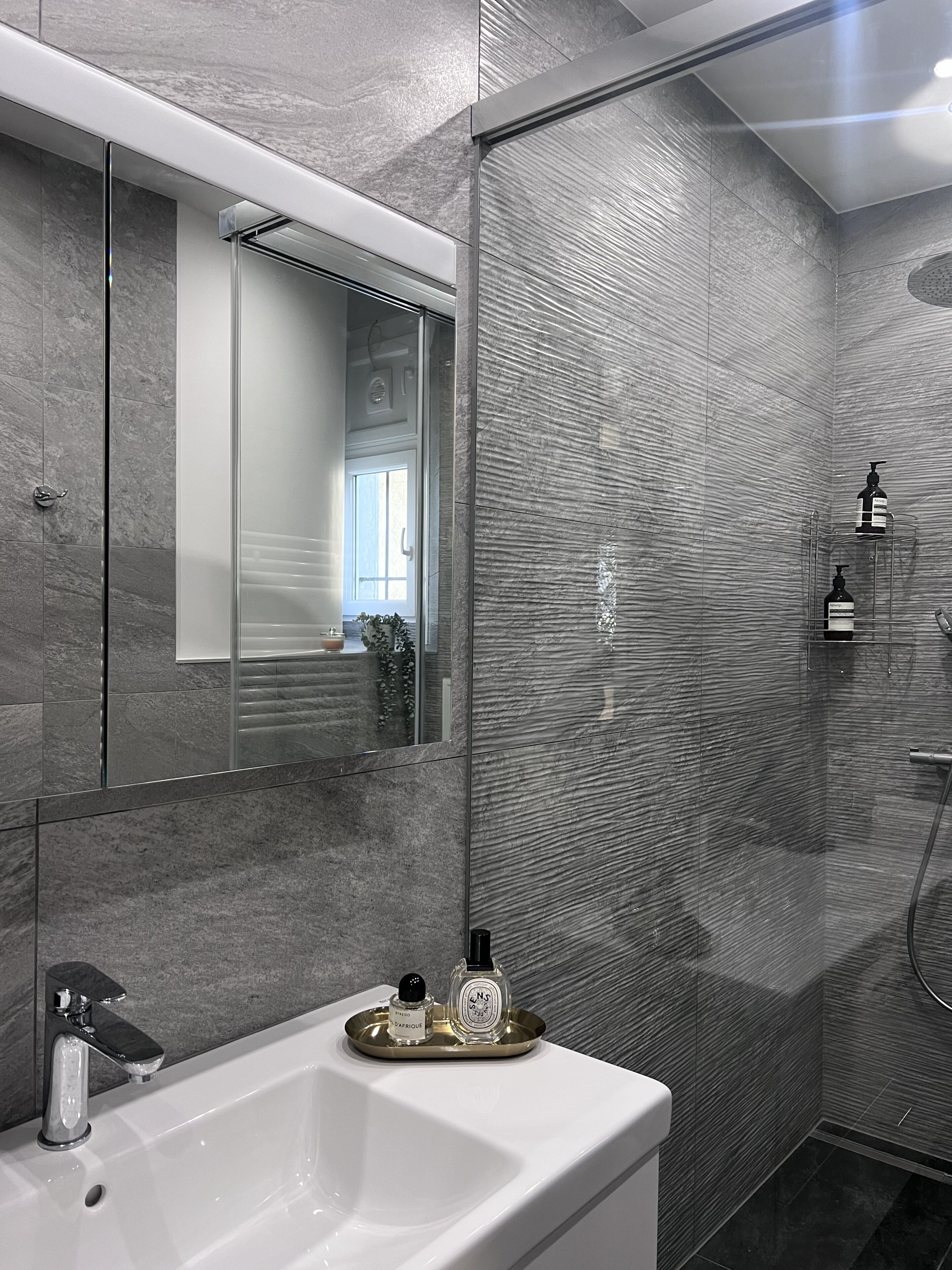 Ein modernes Badezimmer mit einem großen Spiegel, einem weißen Waschbecken und einer Dusche mit strukturierter Wandgestaltung.
