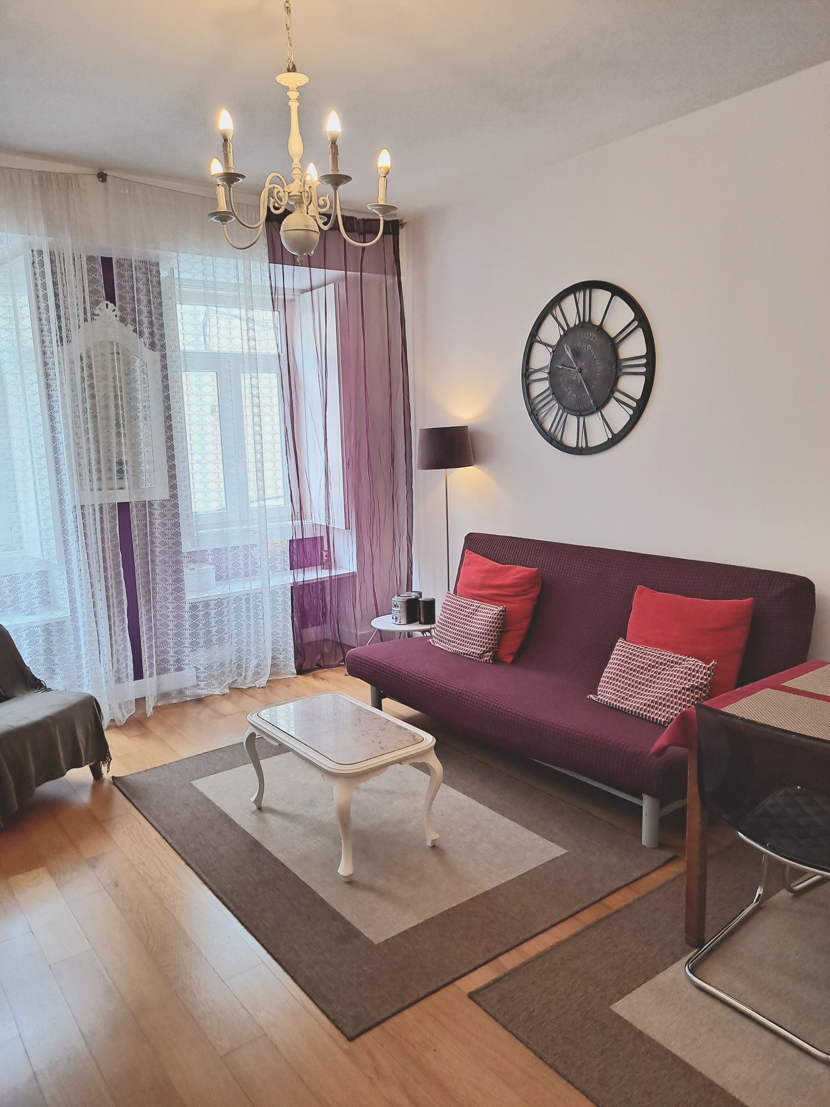 Una sala de estar con sofá, mesa de centro, candelabro, reloj y ventana con cortinas.