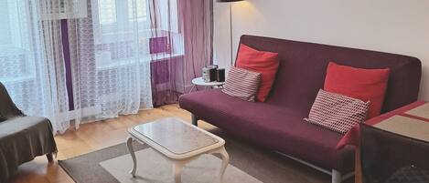 Una sala de estar con sofá, mesa de centro, candelabro, reloj y ventana con cortinas.