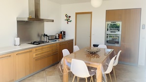 A modern kitchen with wooden cabinets, a stainless steel range hood, and a dining area with a wooden table and white chairs. - 4-star waterfront villa with exceptional sea views (Sari Solenzara)