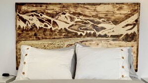 Ein Bett mit weißer Bettwäsche und zwei Kissen, ein Holz-Kopfteil mit einer Berglandschaft und ein Nachttisch mit einer Lampe.