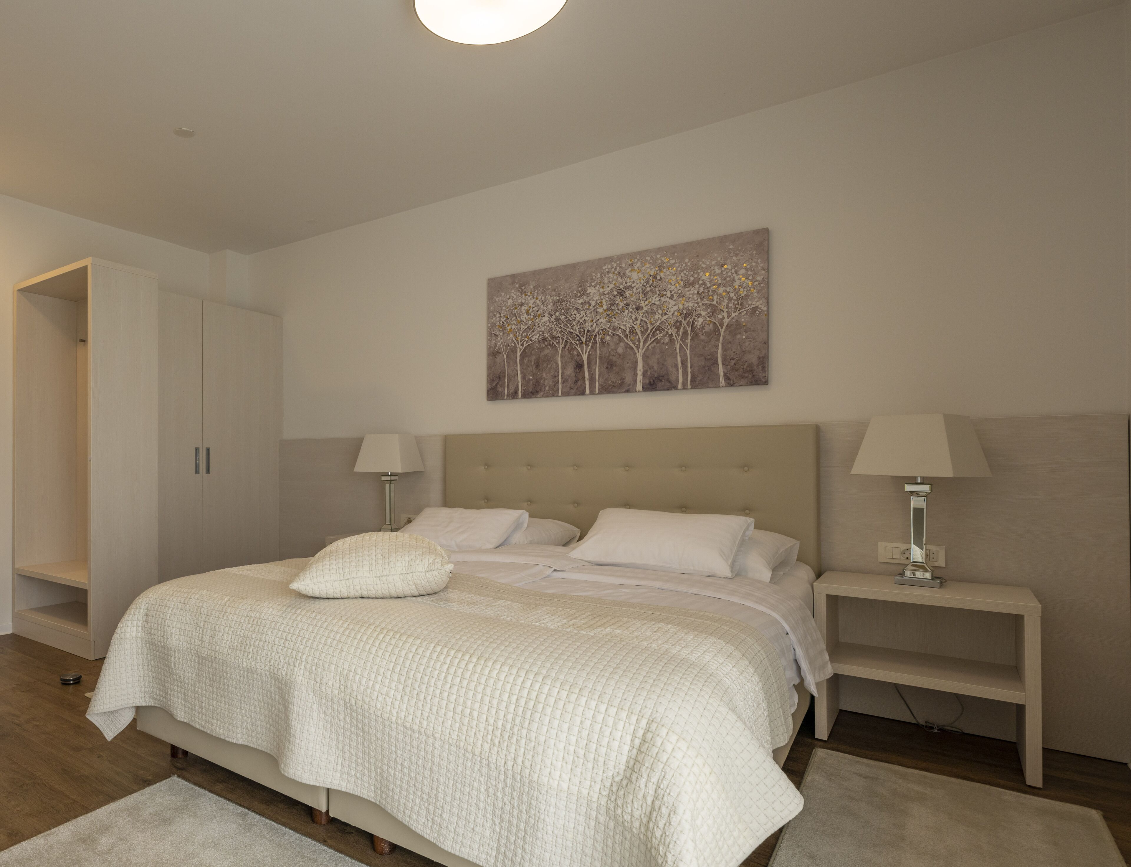 A bedroom with a large bed, two bedside tables, a nightstand, a painting on the wall, and a wardrobe.