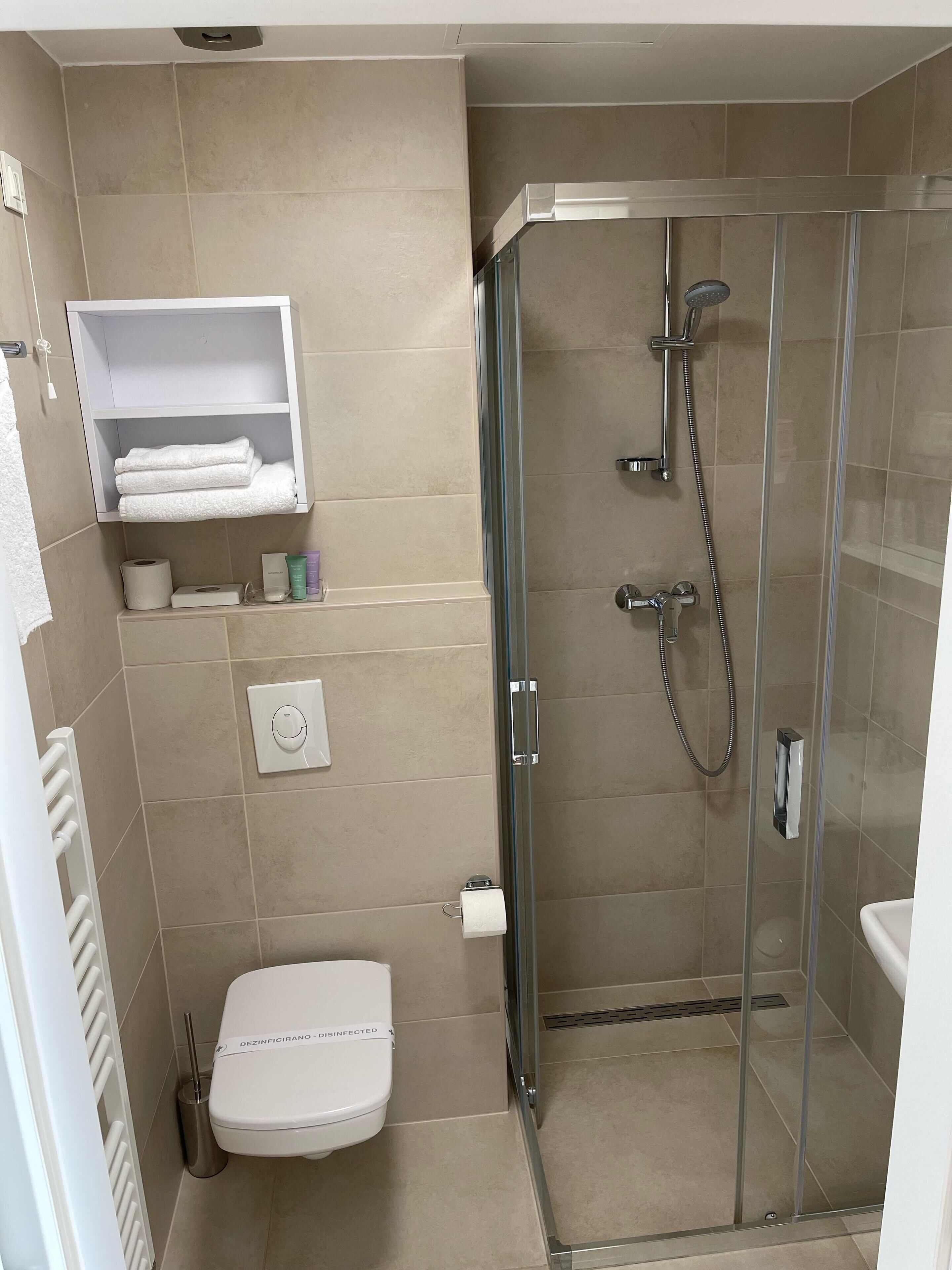 Un baño moderno con ducha de vidrio, inodoro, toallero y estante con artículos de aseo.