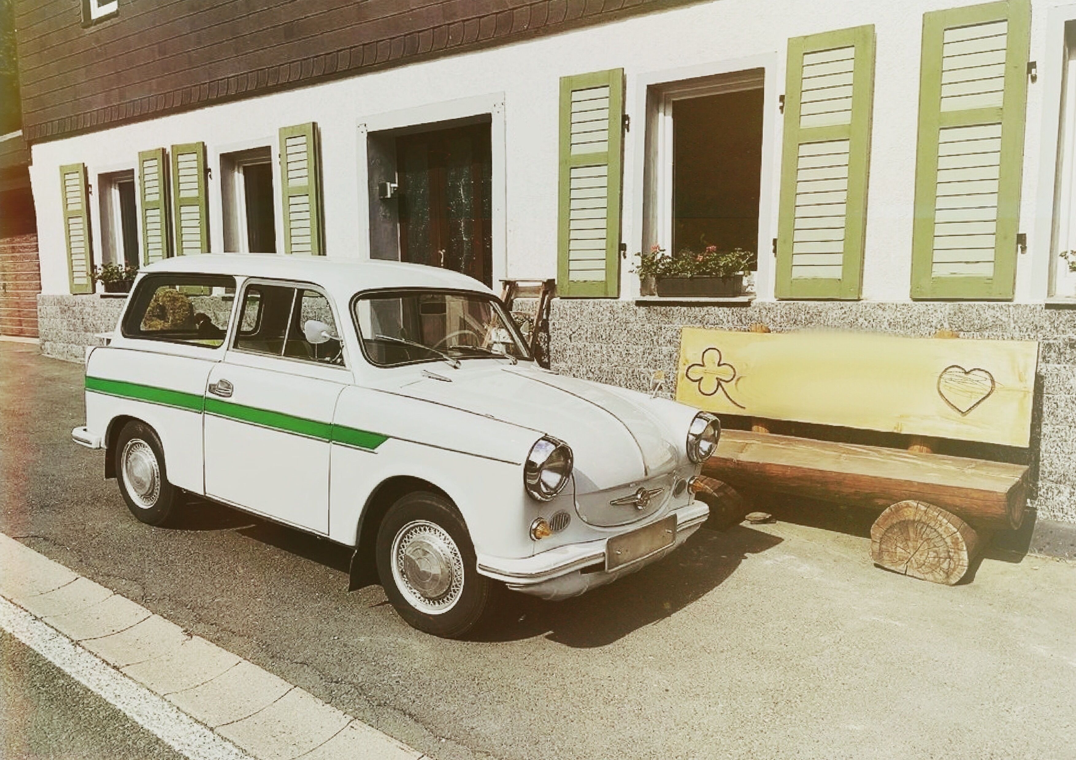 Een klassieke witte auto geparkeerd voor een gebouw met groene luiken en een bank met een hartmotief.