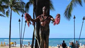 A statue of a man with a surfboard, wearing a lei, on a beach with palm trees and a clear sky. - Waikiki Living, 4 blocks from surf, mountain views, king size bed and office! (Honolulu)