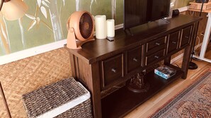 A living room with a dark wooden TV stand, a woven ottoman, a lamp, and a decorative wall with a bamboo pattern. - Waikiki Living, 4 blocks from surf, mountain views, king size bed and office! (Honolulu)