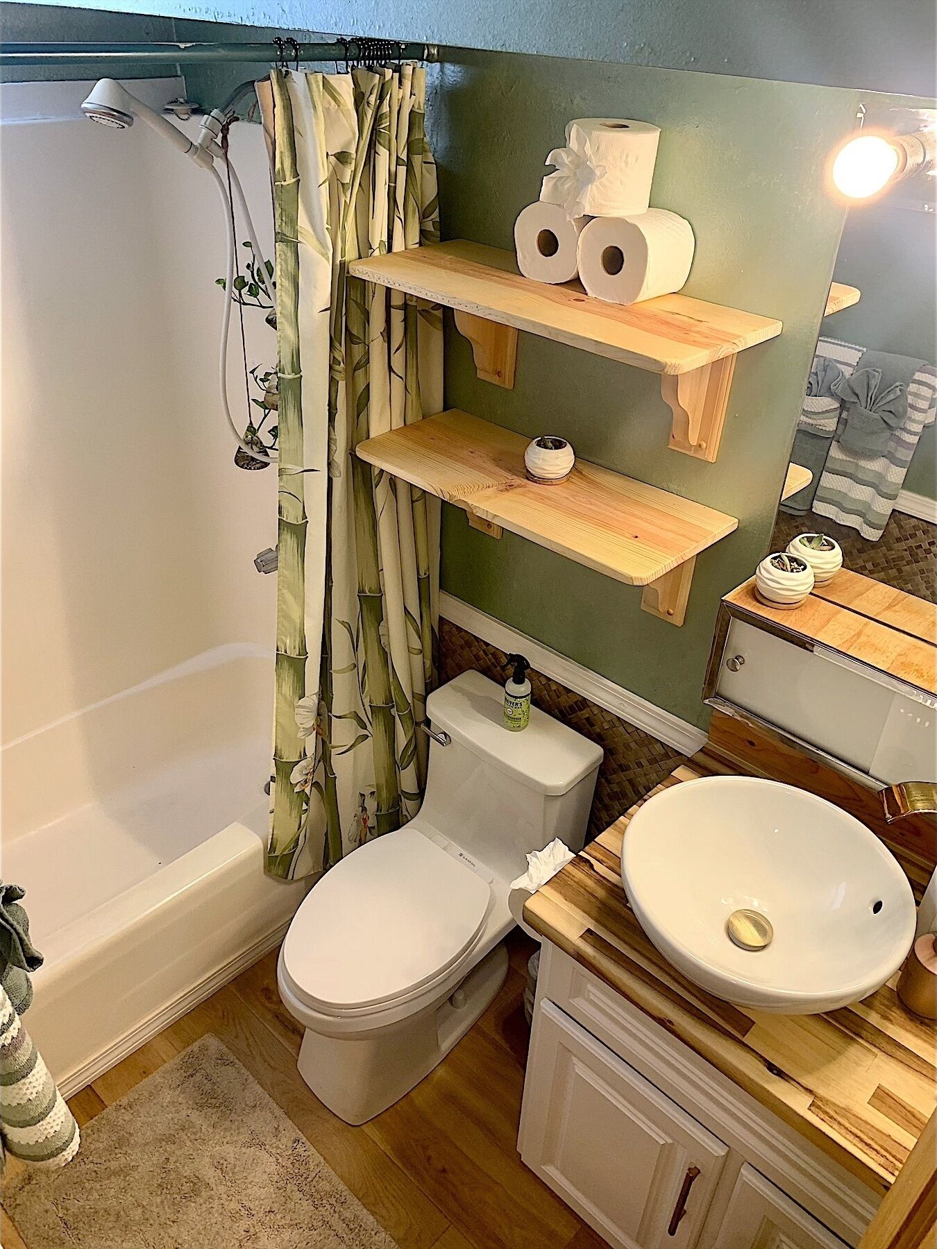 A bathroom with a shower, toilet, and sink. There are wooden shelves above the toilet with toilet paper and a bottle of hand soap.