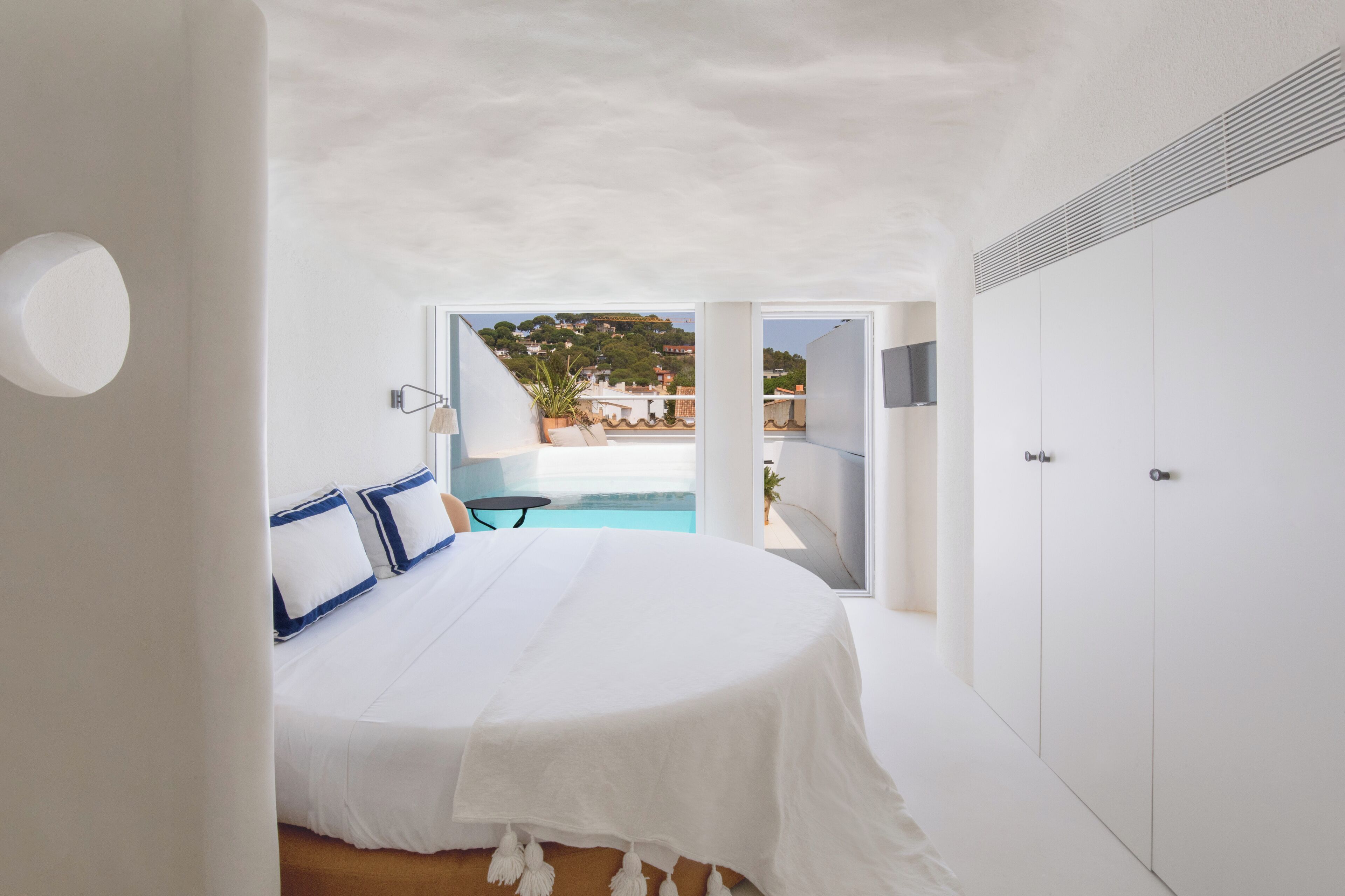 A bedroom with a large bed, a view of a swimming pool, and a balcony with a plant.