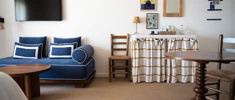 A room with a blue sofa, a wooden chair, a small table, a TV mounted on the wall, and a table with a checkered tablecloth.