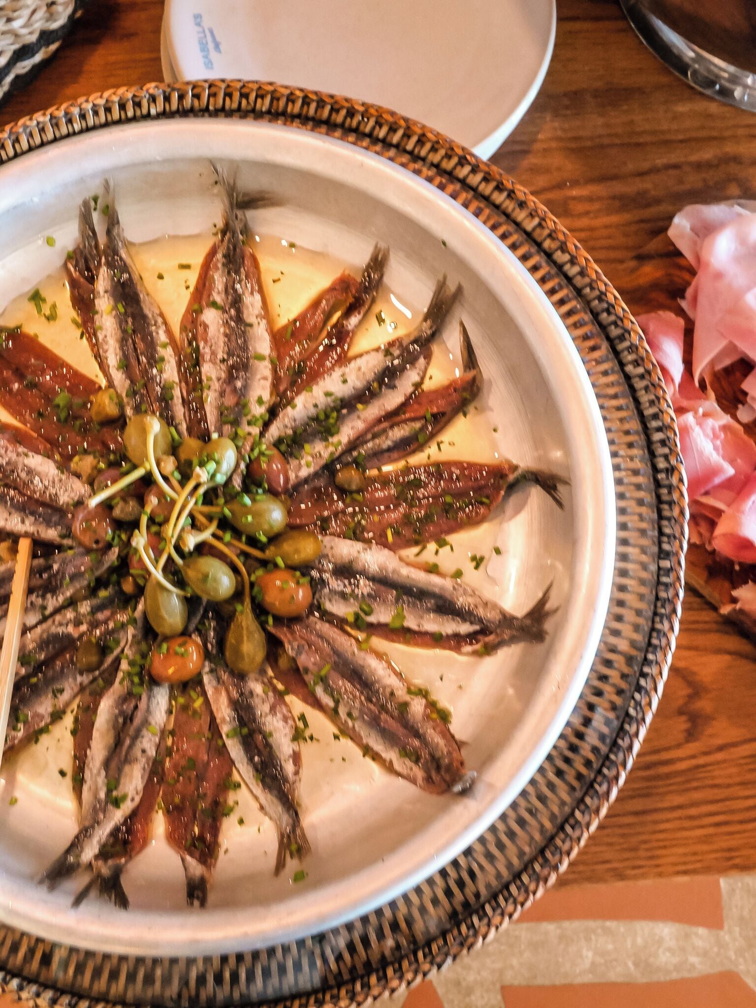 Un plato de sardinas a la parrilla adornado con hierbas y aceitunas, servido en un plato blanco con un borde decorativo.