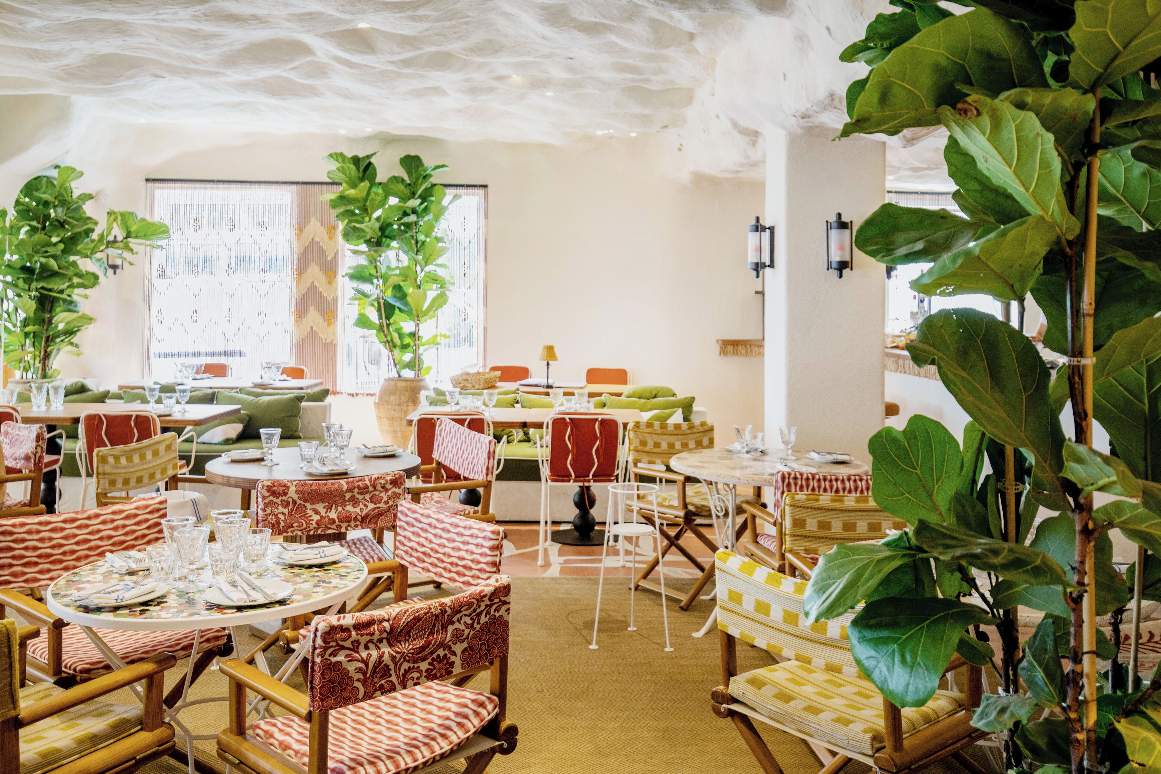 A restaurant with round tables, wicker chairs, and large green plants.