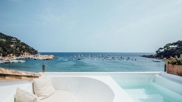 Una piscina infinita moderna con borde blanco, que da a un puerto deportivo con barcos y un paisaje costero.