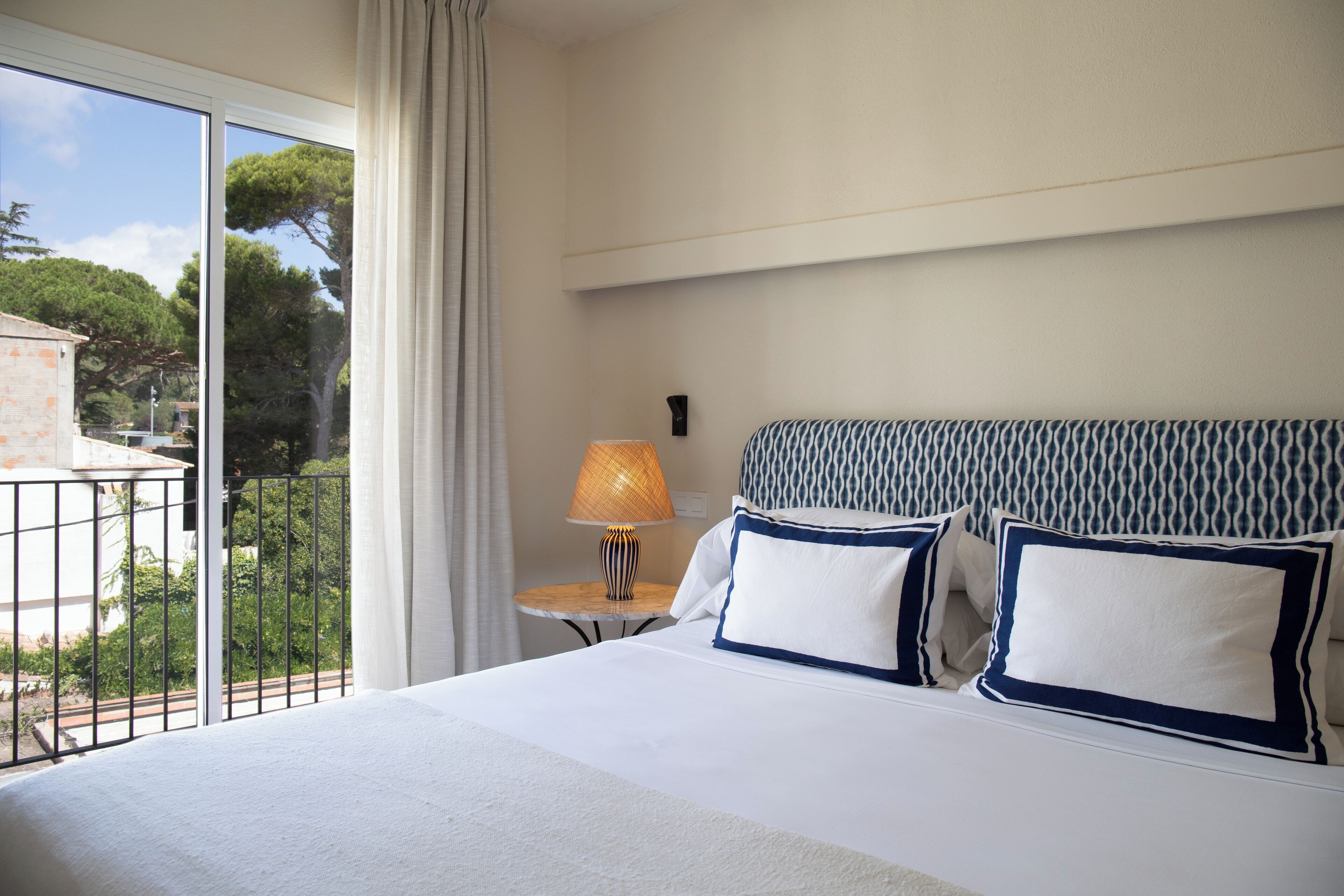 A hotel room with a large bed, a small table with a lamp, and a view of the outside.
