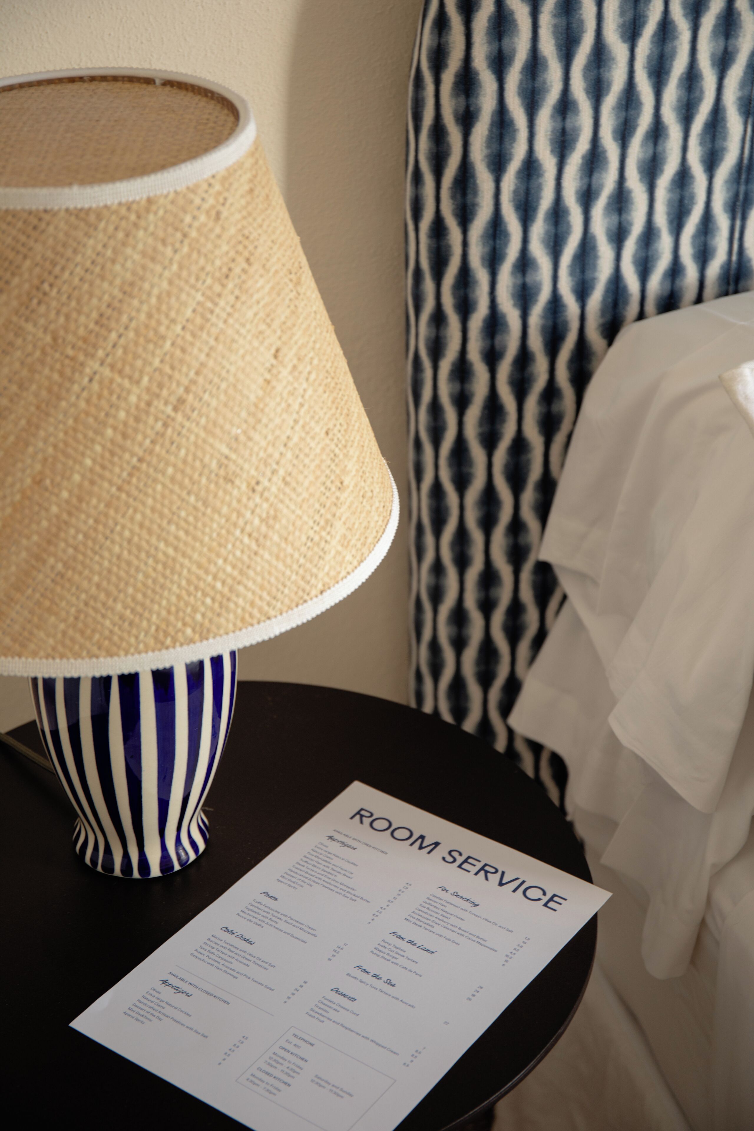 A room service menu on a table with a striped lamp and a patterned curtain in the background.