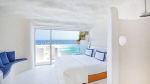 Un dormitorio moderno con una cama grande, una zona de estar con cojines azules, vistas al mar y una mesita con una planta.