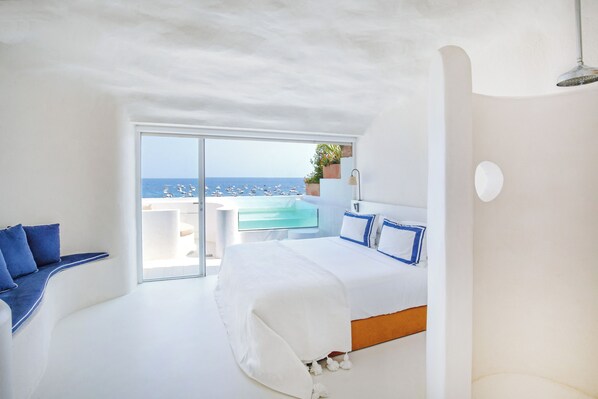 Ein modernes Schlafzimmer mit einem großen Bett, einer Sitzecke mit blauen Kissen, Meerblick und einem kleinen Tisch mit einer Pflanze.