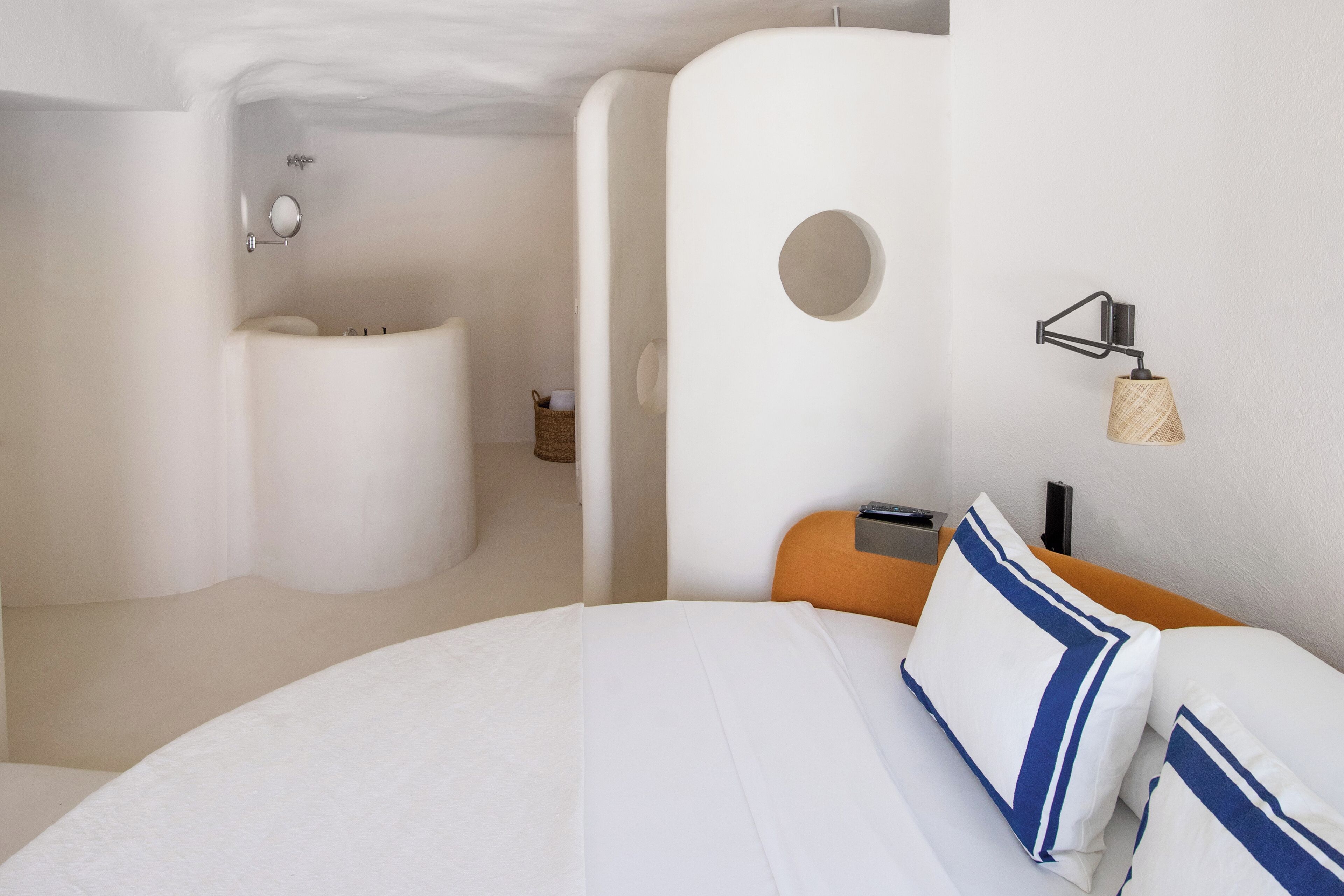 Habitación de hotel moderna con una cama grande, un espejo redondo, un baño pequeño y una lámpara fijada a la pared.