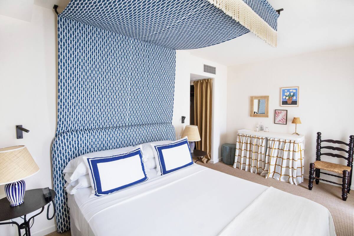 Hotellihuone, jossa on sänky, yöpöytä lampun kanssa, tuoli, pieni pöytä lampun kanssa ja verho.