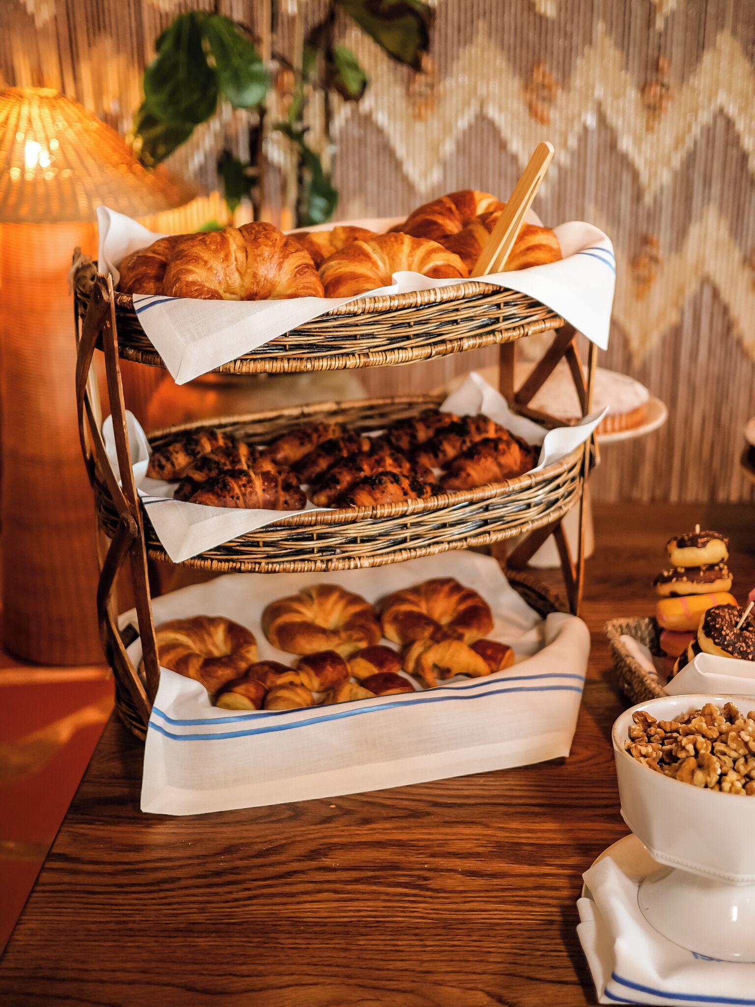 A three-tiered wicker stand with various baked goods, including croissants and muffins, on a wooden table.