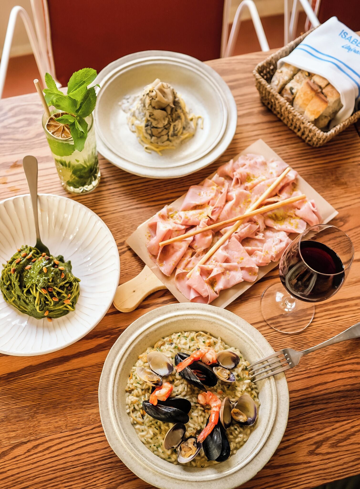 Ein Holztisch mit einer Pasta-Anlage, einer Schüssel Meeresfrüchte-Risotto, einer Platte mit Schinken und einem Glas Rotwein, sowie ein Korb mit Brot.