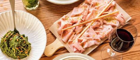 A wooden table with a plate of pasta, a bowl of seafood risotto, a plate of sliced ham, a glass of red wine, and a basket of bread.